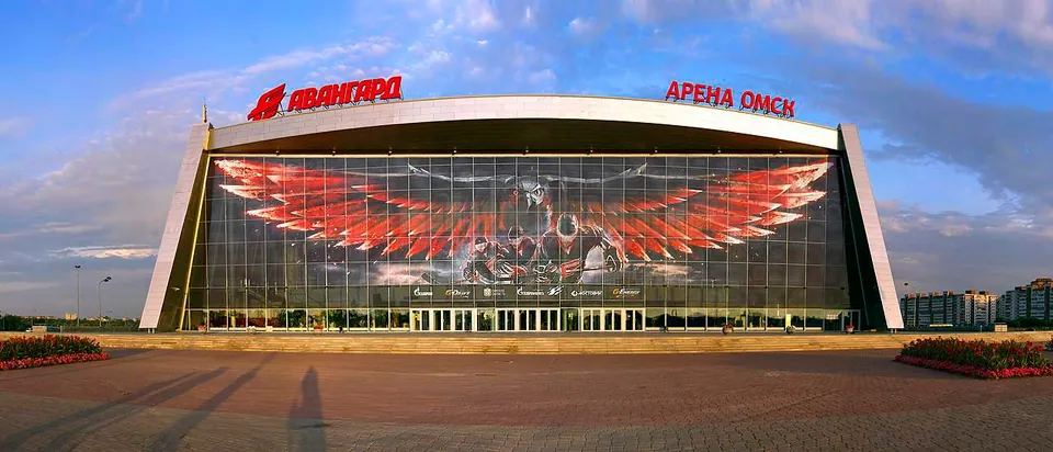 อาคารขนาดใหญ่มีป้ายสีแดงเขียนว่า 'АВАНГАРД' และ 'АРЕНА ОМСК' ด้านหน้าเป็นกระจกมีภาพปีกสีแดงขนาดใหญ่
