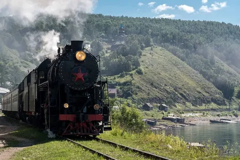 รถไฟหัวจักรสีดำพ่นควันสีขาววิ่งอยู่บนรางรถไฟริมแม่น้ำ โดยมีเนินเขาปกคลุมด้วยต้นไม้และอาคารหลังหนึ่งตั้งอยู่บนยอดเขาเป็นฉากหลัง