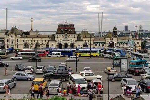อาคารสถานีรถไฟสไตล์ยุโรปขนาดใหญ่ตั้งอยู่หลังลานจอดรถที่มีรถยนต์และรถบัสหลายสีจอดเรียงราย โดยมีผู้คนเดินอยู่บริเวณหน้าอาคาร
