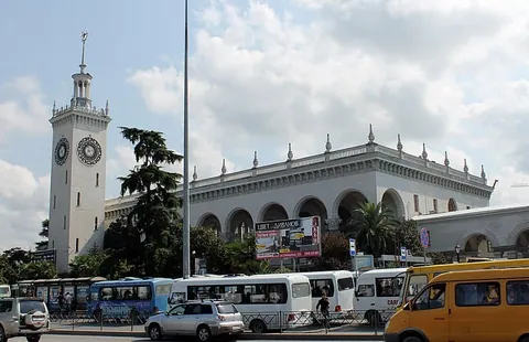 อาคารสถานีรถไฟสีขาวทรงยาวมีซุ้มประตูโค้งเรียงกัน มีหอคอยนาฬิกาสูงตั้งอยู่ด้านซ้าย และมีรถบัสหลายสีจอดเรียงรายอยู่ด้านหน้า