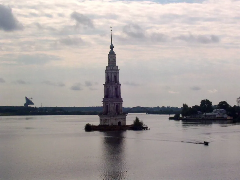 หอคอยระฆัง Kalyazin สีชมพูตั้งอยู่บนเกาะกลางแม่น้ำที่มีเรือเล็กแล่นผ่าน ภายใต้ท้องฟ้าที่มีเมฆปกคลุม