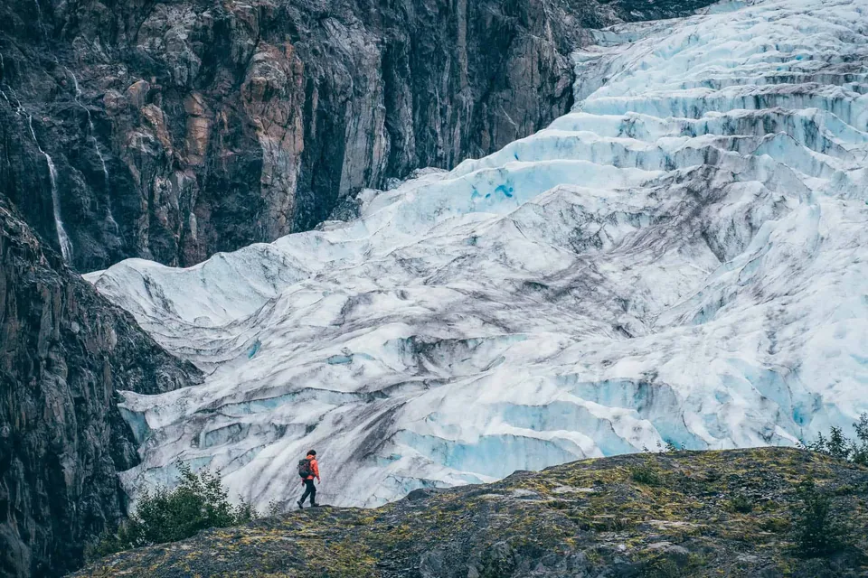 ธารน้ำแข็ง Exit Glacier สีขาวอมฟ้าไหลลงจากหน้าผาหิน มีคนใส่เสื้อสีส้มเดินอยู่บนพื้นหินด้านหน้า