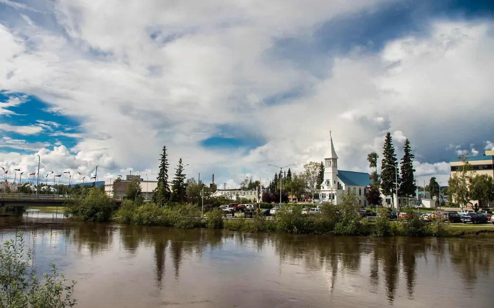 ภาพเมืองแฟรร์แบงก์ส (Fairbanks) แสดงแม่น้ำที่ทอดยาวผ่านพื้นที่สีเขียว มีอาคารสีขาวหลังคาแหลมตั้งอยู่ท่ามกลางต้นไม้สูง และท้องฟ้าเต็มไปด้วยเมฆ