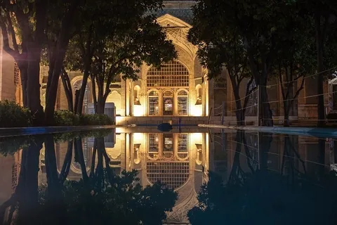 สวนของสุสานกวีฮาเฟซในชีราซ แสดงให้เห็นอาคารที่มีแสงไฟส่องสว่างและสะท้อนลงบนผิวน้ำโดยมีต้นไม้ใหญ่เรียงรายอยู่สองข้างทาง
