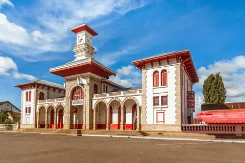 อาคารสถานีรถไฟสีขาวและสีแดงที่มีหอคอยสูง มีป้ายเขียนว่า ANTISIRABE ตั้งอยู่ใต้ท้องฟ้าสีฟ้าที่มีเมฆ