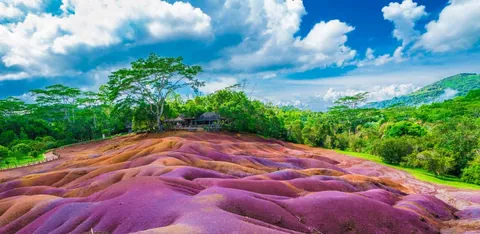 เนินดินที่มีพื้นผิวเป็นลอนคลื่นในเฉดสีม่วง ชมพู และส้ม โอบล้อมด้วยต้นไม้สีเขียวและท้องฟ้าสีน้ำเงินที่มีเมฆขาว