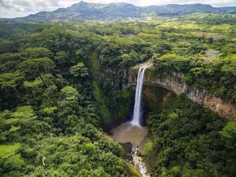ภาพมุมสูงของน้ำตกซามาเรล (Chamarel Falls) ที่ไหลลงจากหน้าผาสูงท่ามกลางป่าไม้เขียวขจีและภูเขาในเบื้องหลัง