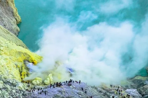ภาพมุมสูงของแอ่งน้ำสีฟ้าอมเขียวที่มีกลุ่มควันสีขาวหนาปกคลุมบริเวณขอบแอ่ง ซึ่งพื้นดินโดยรอบมีสีเหลืองและเทา มีกลุ่มคนจำนวนมากยืนกระจายอยู่บนพื้นหินบริเวณขอบแอ่ง
