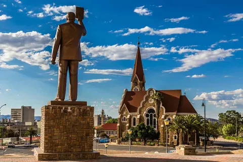 รูปปั้นบุรุษยืนถือหนังสือบนแท่นหินด้านหน้าโบสถ์ Christuskirche ที่มีหลังคาสีแดงและหอคอยแหลม ภายใต้ท้องฟ้าสีฟ้า