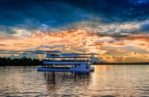 เรือสีขาวสองชั้นลอยอยู่บนผิวน้ำที่มีแสงสีส้มและฟ้าสะท้อนอยู่ โดยมีกลุ่มคนยืนอยู่บนดาดฟ้าชั้นบน และแนวป่าไม้ทอดยาวอยู่ตามแนวขอบฟ้า