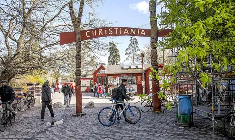 ป้ายไม้สีแดงขนาดใหญ่เขียนคำว่า CHRISTIANIA ตั้งอยู่เหนือทางเดินหินกรวด มีผู้คนเดินและปั่นจักรยานผ่านหน้าอาคารไม้สีแดงหลังเล็ก