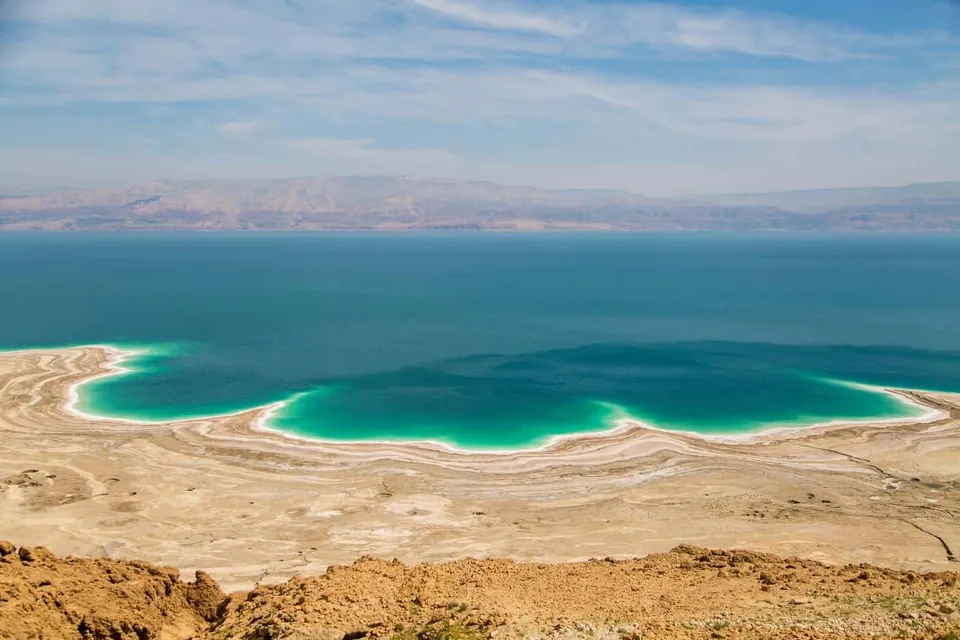 ภาพมุมสูงของทะเลสาบเดดซี (Dead Sea) ที่ขอบน้ำมีสีฟ้าอมเขียวตัดกับผืนทรายสีน้ำตาล และแนวภูเขาไกลๆ ใต้ท้องฟ้าสีฟ้า