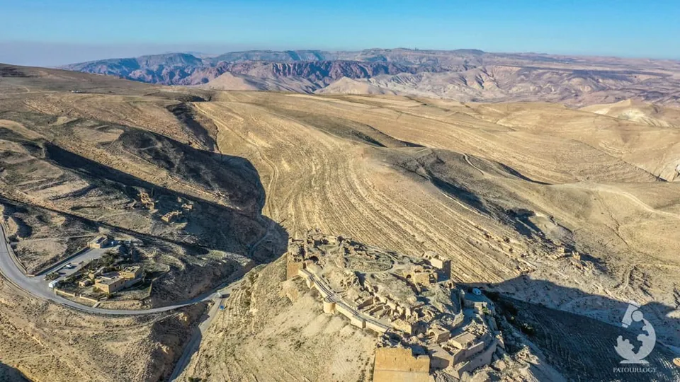 ภาพมุมสูงของปราสาทโชบาค (Shobak) ตั้งอยู่บนเนินเขาหินสีน้ำตาลในภูมิประเทศแห้งแล้ง มีทางคดเคี้ยวและอาคารขนาดเล็กอยู่ใกล้กัน