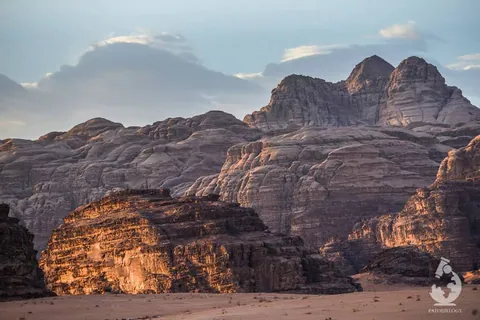 ทิวทัศน์ทะเลทรายวาดิรัม (Wadi Rum) แสดงหน้าผาหินขนาดใหญ่ที่มีชั้นหินซ้อนทับกันเป็นชั้น ๆ โผล่ขึ้นมาจากพื้นทรายสีอ่อน ภายใต้ท้องฟ้าสีฟ้าที่มีเมฆบางเบา