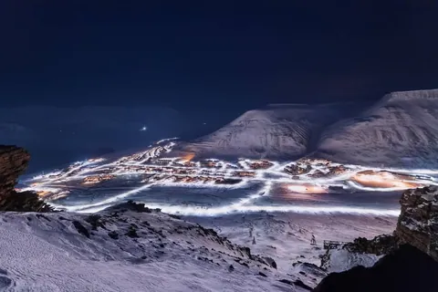 ภาพเมืองลองเยียร์เบียน (Longyearbyen) ที่ปกคลุมด้วยหิมะในเวลากลางคืน มีแสงไฟจากอาคารและถนนสว่างตัดกับทิวเขาและท้องฟ้ามืด