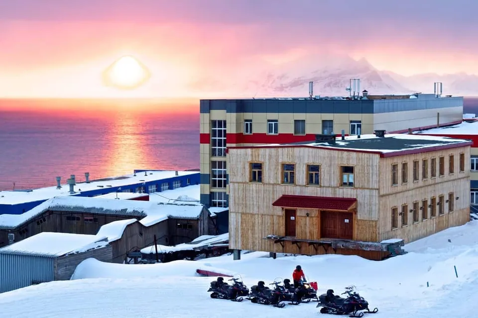 ภาพเมือง Barentsburg ที่ปกคลุมด้วยหิมะ มีอาคารไม้และคอนกรีตตั้งอยู่ริมทะเล พร้อมรถสโนว์โมบิลจอดเรียงกันหน้าอาคาร