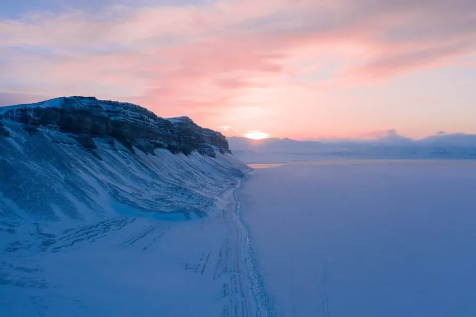 ทิวทัศน์หุบเขา Longyeardalen ที่ปกคลุมด้วยหิมะและน้ำแข็ง มีหน้าผาหินด้านซ้าย และท้องฟ้าสีชมพูอมม่วงจากแสงอาทิตย์ที่ขอบฟ้า