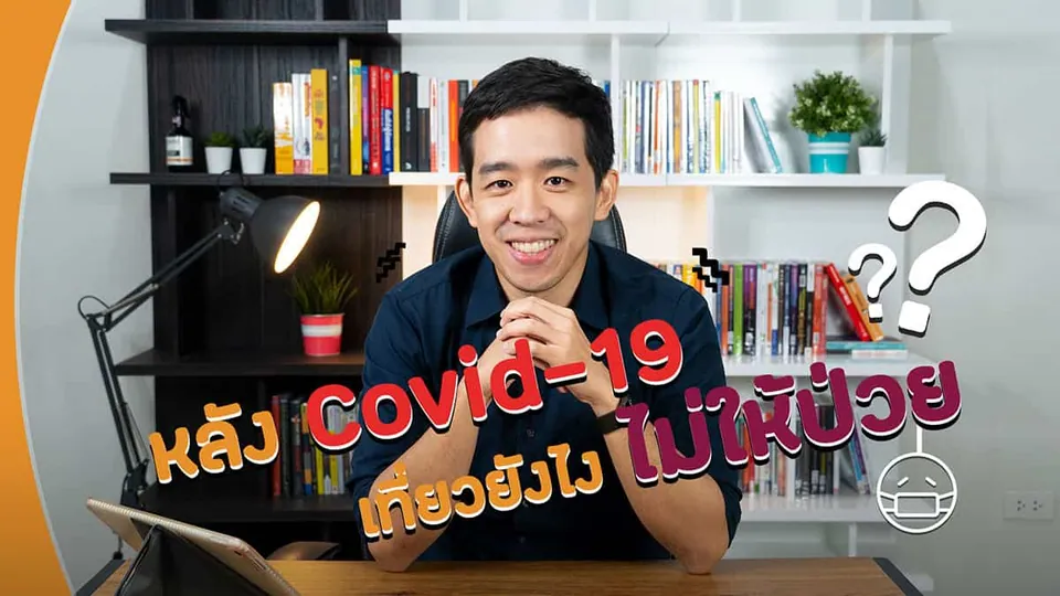ชายในเสื้อเชิ้ตสีน้ำเงินนั่งยิ้มที่โต๊ะทำงานหน้าชั้นวางหนังสือ มีโคมไฟตั้งโต๊ะและข้อความ "Kลวง Covid-19" พร้อมเครื่องหมายคำถามปรากฏบนภาพ