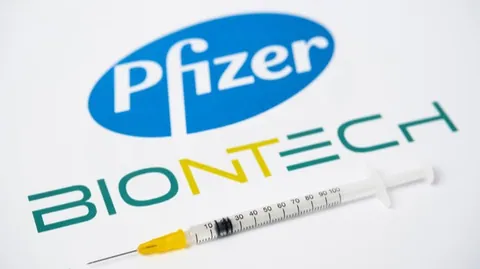 ภาพโลโก้ Pfizer สีน้ำเงินและโลโก้ BioNTech สีเขียว-เหลือง วางอยู่บนพื้นหลังสีขาว โดยมีหลอดฉีดยาที่มีเข็มและส่วนปลายสีเหลืองวางพาดอยู่ด้านหน้า