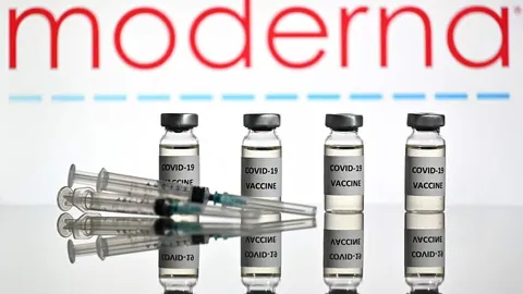 ภาพขวดวัคซีนสี่ขวดที่มีฉลากเขียนว่า COVID-19 VACCINE วางเรียงกันบนพื้นผิวสะท้อนแสง มีเข็มฉีดยาวางอยู่ด้านหน้า และโลโก้คำว่า moderna สีแดงปรากฏอยู่ด้านบน