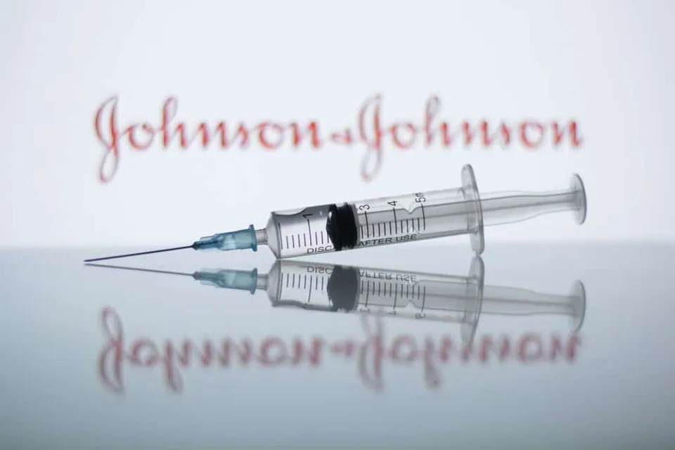 ภาพหลอดฉีดยาพร้อมเข็มวางอยู่บนพื้นผิวสะท้อนแสง มีตัวอักษร Johnson & Johnson สีแดงปรากฏอยู่ด้านหลัง