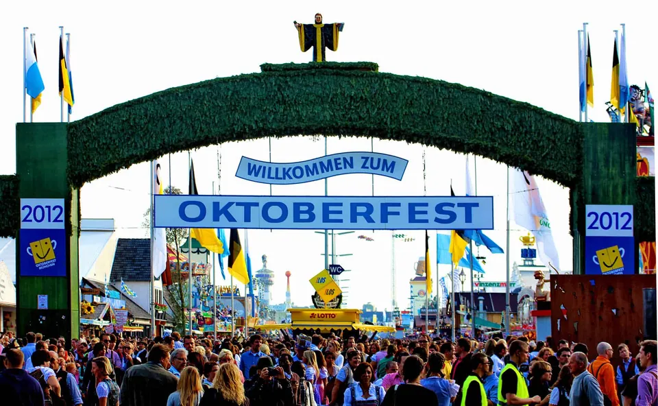 ประตูทางเข้าขนาดใหญ่ประดับด้วยพืชสีเขียว มีป้ายสีน้ำเงินเขียนว่า WILLKOMMEN ZUM OKTOBERFEST และตัวเลข 2012 อยู่ด้านข้าง ท่ามกลางฝูงชนจำนวนมาก
