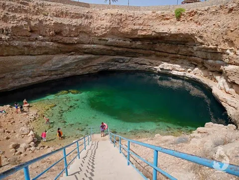 บ่อ Bimmah sinkhole น้ำสีเขียวมรกตอยู่ภายในแอ่งหินสีน้ำตาล มีบันไดคอนกรีตพร้อมราวกั้นสีน้ำเงินทอดลงไปยังขอบน้ำ และมีผู้คนยืนอยู่บริเวณริมตลิ่ง