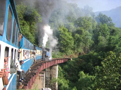รถไฟสีน้ำเงินและขาวกำลังวิ่งบนสะพานเหล็กสีแดงท่ามกลางป่าไม้เขียวขจี โดยมีควันสีขาวพวยพุ่งออกมาจากหัวรถจักร
