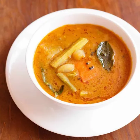 ชามซุปซัมบาร์ (Sambar) สีส้มในชามสีขาว มีถั่วและผักชิ้นใหญ่ลอยอยู่ วางบนจานรองสีขาว