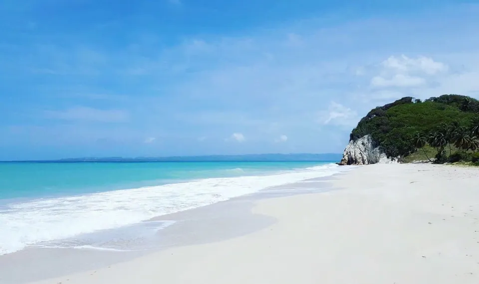 หาดทรายสีขาวทอดยาวติดกับทะเลสีฟ้าอมเขียว มีคลื่นซัดเข้าฝั่ง และมีเนินเขาปกคลุมด้วยต้นไม้สีเขียวตั้งอยู่ด้านขวา