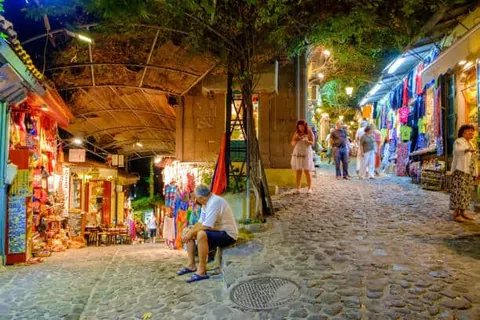 ถนนปูหินในเวลากลางคืนที่มีผู้คนเดินอยู่สองข้างทาง ซึ่งเต็มไปด้วยร้านค้าที่ประดับไฟสว่างไสว