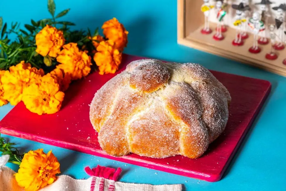 ขนมปัง Pan de ánimas ทรงกลมสีน้ำตาลทองโรยน้ำตาล วางบนจานสีแดง มีดอกดาวเรืองสีส้มและกล่องตุ๊กตาอยู่ข้างหลัง