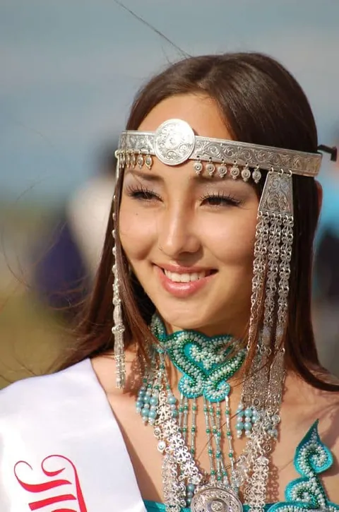 หญิงผมยาวสวมเครื่องประดับศีรษะและสร้อยคอสีเงินประดับลูกปัดสีเขียว พร้อมสายสะพายสีขาวมีตัวอักษรสีแดง