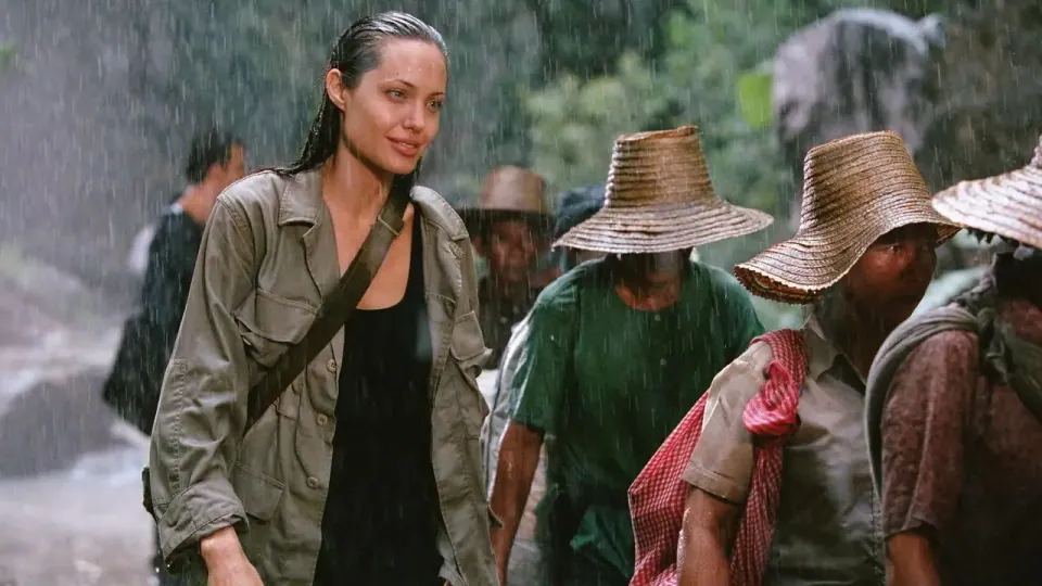 หญิงผมเปียเปียฝนสวมเสื้อแจ็คเก็ตสีเทาและสายสะพายคาดหน้าอก ยืนอยู่ด้านหน้ากลุ่มคนสวมหมวกฟางในสภาพฝนตก