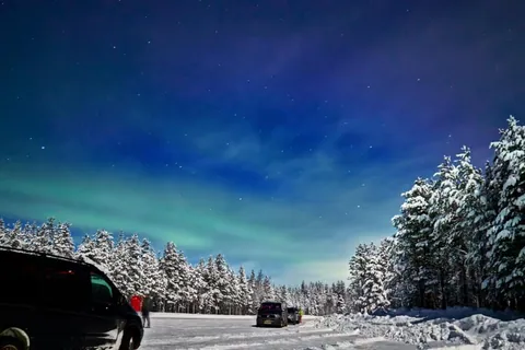 ภาพทิวทัศน์ยามค่ำคืนที่มีแสงสีเขียวอมฟ้าปรากฏบนท้องฟ้าเหนือป่าสนที่ปกคลุมด้วยหิมะ โดยมีรถยนต์จอดอยู่บนพื้นหิมะด้านหน้า