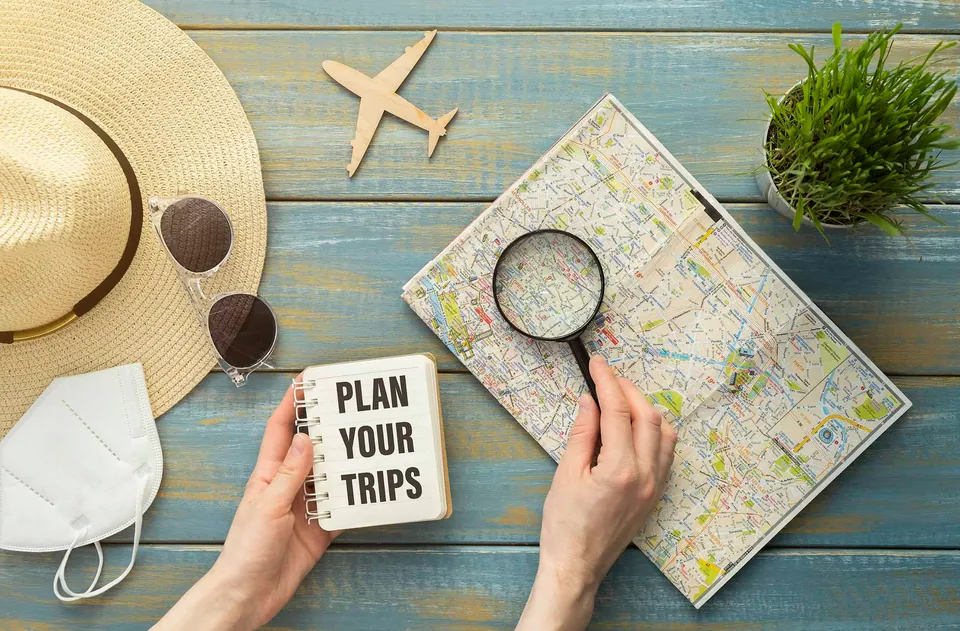 มือทั้งสองข้างถือสมุดบันทึกที่มีข้อความ "PLAN YOUR TRIPS" และใช้แว่นขยายส่องแผนที่บนโต๊ะไม้สีน้ำเงิน พร้อมหมวกฟาง แว่นกันแดด หน้ากากอนามัย และโมเดลเครื่องบิน