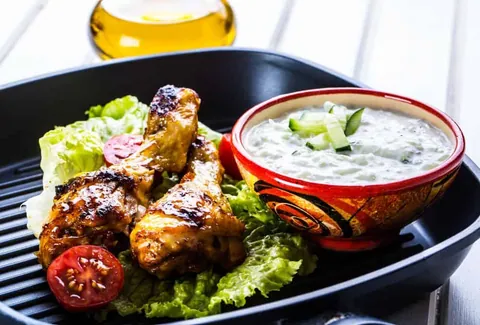 จานอาหารสีดำมีไก่ย่างชิ้นใหญ่บนผักกาดหอมและมะเขือเทศหั่นครึ่ง พร้อมชามซอสครีมสีขาวมีแตงกวาหั่นเป็นชิ้นเล็ก ๆ วางข้างกัน