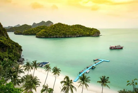 ทิวทัศน์หาดทรายขาวเรียงรายด้วยต้นมะพร้าว มีเกาะปกคลุมด้วยป่าเขียวขจีทอดยาวในน้ำทะเลสีฟ้าอมเขียว พร้อมเรือลำหนึ่งลอยอยู่กลางทะเลและแพสีน้ำเงินทอดยาวจากฝั่ง