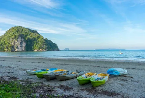 เรือคายัคหลายลำหลากสีสันจอดอยู่บนหาดทราย มีภูเขาหินปกคลุมด้วยต้นไม้สีเขียวตั้งอยู่ริมทะเลเบื้องหลัง