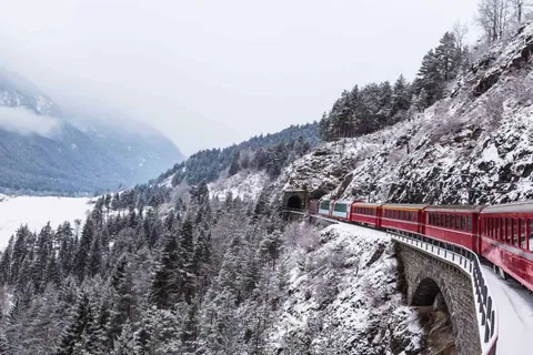 ขบวนรถไฟสีแดงวิ่งผ่านสะพานโค้งหินบนทางรถไฟที่ขดตัวไปตามไหล่เขาที่ปกคลุมด้วยหิมะและป่าสน