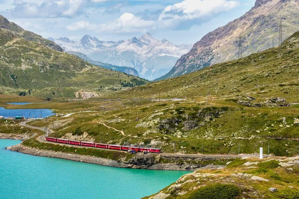 ขบวนรถไฟสีแดงแล่นไปตามรางที่คดเคี้ยวริมทะเลสาบสีฟ้าอมเขียว ท่ามกลางเนินเขาหญ้าเขียวขจีและยอดเขาหินในเบื้องหลัง