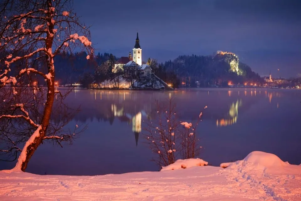 ภาพทิวทัศน์ยามค่ำของเกาะกลางน้ำที่มีอาคารสีขาวหลังคาแหลมและหอคอยตั้งอยู่ สะท้อนเงาบนผิวน้ำ ริมตลิ่งด้านหน้ามีต้นไม้และพุ่มไม้ปกคลุมด้วยหิมะ