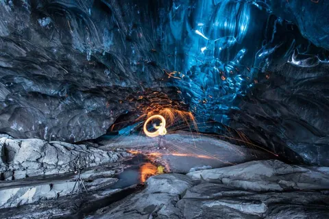 ภาพภายในถ้ำที่มีผนังหินสีน้ำเงินเข้มและพื้นหินสีเทา มีคนยืนอยู่ตรงกลางกำลังสร้างวงแหวนแสงสีส้มจากประกายไฟ