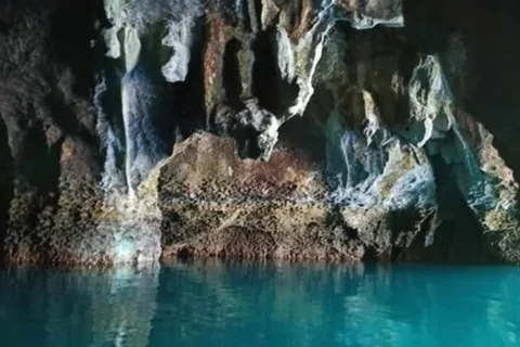 ภาพภายในถ้ำที่มีผนังหินสีน้ำตาลและเทาตั้งอยู่เหนือผิวน้ำสีฟ้าอมเขียวที่สะท้อนแสง