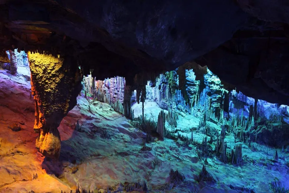 ภาพภายในถ้ำที่มีเสาหินและ stalactites ยื่นลงมาจากเพดาน ถูกส่องสว่างด้วยแสงสีเหลือง ส้ม และน้ำเงิน
