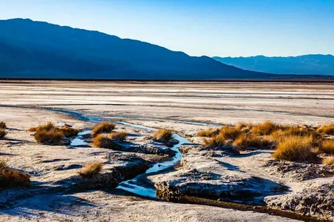 ภาพทิวทัศน์ของพื้นที่ราบโล่งกว้างที่มีลำธารน้ำเล็กๆ ไหลคดเคี้ยวผ่านพุ่มหญ้าสีน้ำตาลแห้งแล้ง โดยมีแนวเทือกเขาสูงทอดยาวเป็นฉากหลังภายใต้ท้องฟ้าสีฟ้าใส