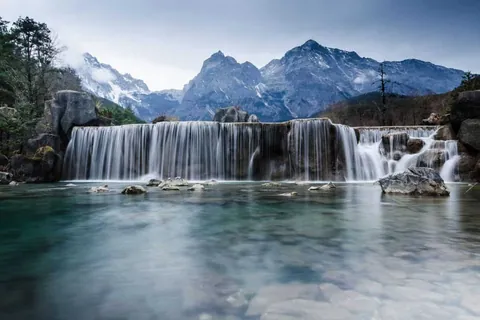 ภาพน้ำตกไหลลงสู่แอ่งน้ำใสสีฟ้า มีก้อนหินขนาดใหญ่กระจายอยู่รอบๆ และภูเขาสูงตั้งตระหง่านเป็นฉากหลัง
