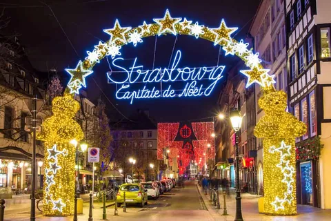 ประตูซุ้มไฟรูปดาวสีเหลืองและขาว มีป้ายไฟสีฟ้าเขียนว่า 'Strasbourg Capitale de Noël' ตั้งอยู่หน้าถนนที่มีอาคารและรถยนต์จอดอยู่