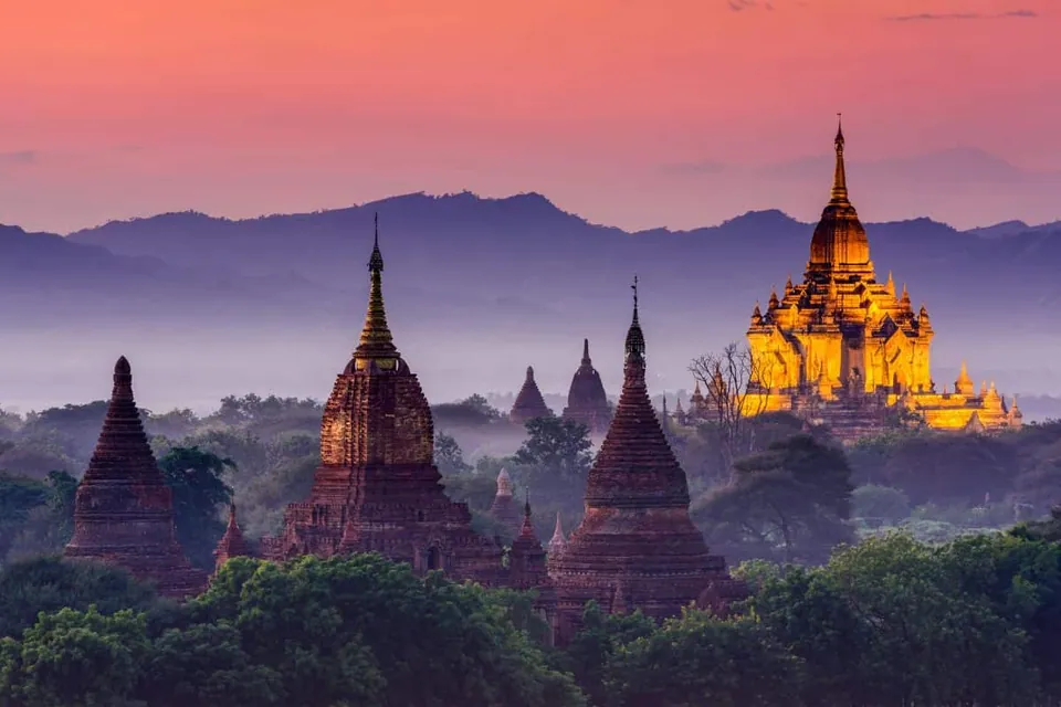 ภาพพระเจดีย์หลายหลังตั้งอยู่ท่ามกลางต้นไม้ มีพระเจดีย์หลังหนึ่งส่องแสงสีทองอยู่ด้านขวา ใต้ท้องฟ้าสีชมพูอมม่วงและแนวภูเขาไกลๆ