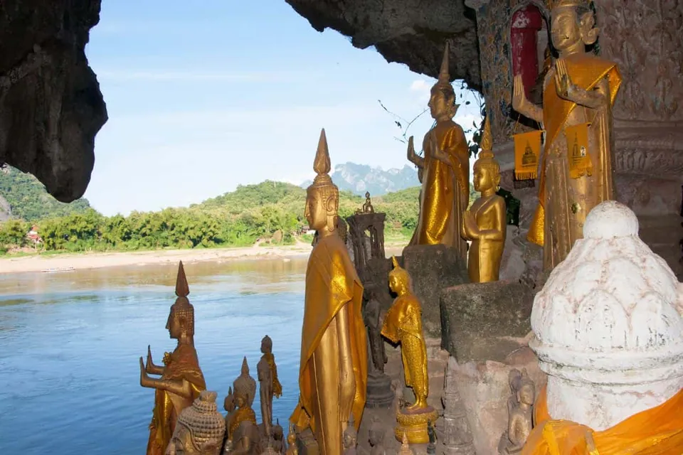 รูปปั้นพระพุทธรูปสีทองหลายองค์ตั้งเรียงรายอยู่ภายในถ้ำหิน มองเห็นแม่น้ำสายกว้างและทิวเขาเขียวขจีอยู่เบื้องหลัง